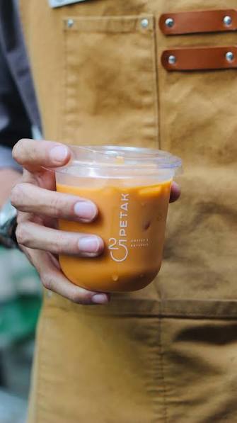 Tangan bercelemek cokelat memegang gelas es kopi susu berlogo PETAK 25