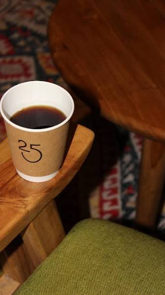 Segelas kopi hitam dalam cangkir kertas kraft di atas sandaran kursi kayu, dengan karpet bermotif di bawahnya